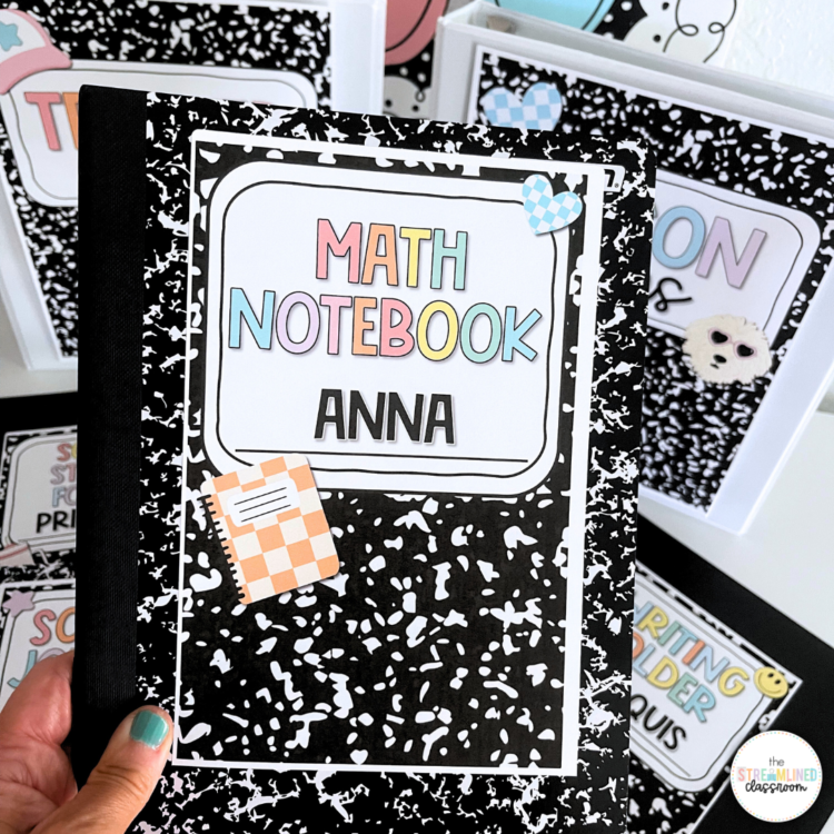 A black-and-white composition notebook with an editable label that reads "Math Notebook" in pastel-colored letters and "Anna" in bold black text. The label features cute clipart, including a small orange-and-white checkered notebook and blue heart-shaped accents. In the background, other labeled binders and notebooks are visible, featuring the same composition notebook theme.