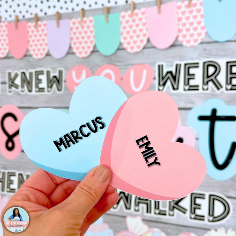 A teacher holds two pastel pink and blue candy heart cutouts with editable text fields, personalized with student names. The background showcases the completed Valentine’s bulletin board, giving a close-up look at the cute classroom decor details.