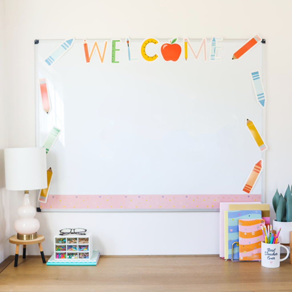 A bright and organized classroom setup featuring a whiteboard with a 'WELCOME' banner in colorful, playful font with school-themed illustrations like pencils and an apple. The whiteboard is decorated with cutouts of colorful pencils along the edges and a pastel pink border with small stars. Below the board, a wooden desk holds a small white lamp with a wooden base, a glasses case, a stationery organizer filled with colorful supplies, and a mug labeled 'Best Teacher Ever' holding pens and pencils. To the right, neatly stacked notebooks and folders in pastel colors add to the cheerful and organized aesthetic.