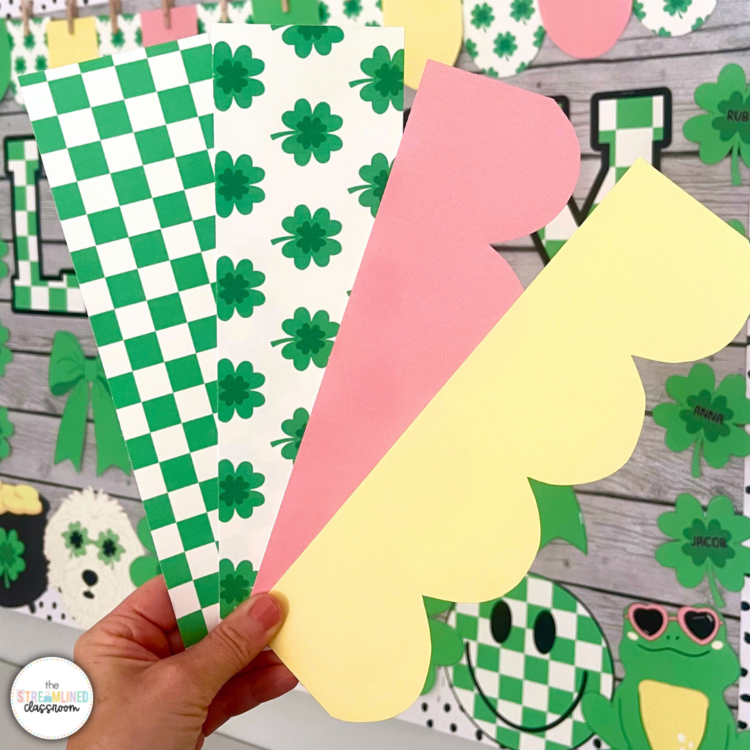 Close-up photo of a hand holding four printed border pieces with green checkerboard, shamrock print, pastel pink, and pastel yellow designs, set against the Lucky Vibes bulletin board display.