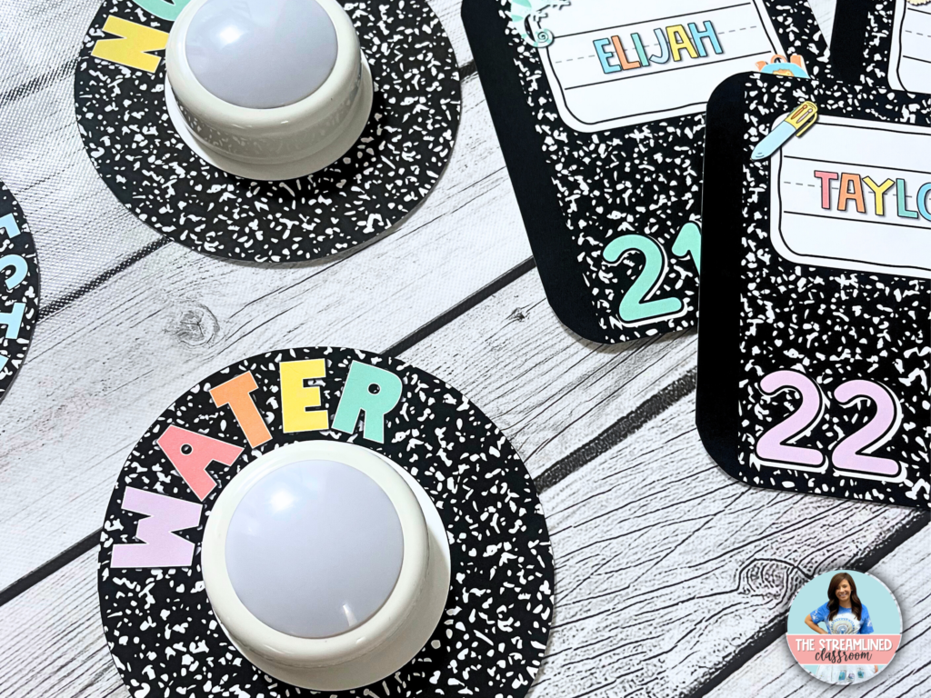 A set of composition notebook-themed push light hall passes displayed on a wooden surface. Each pass has bold, pastel-colored text indicating different hall pass uses, such as "Water." Matching student desk name tags with colorful labels and numbers are also visible, making this an ideal addition to a cohesive classroom decor theme.