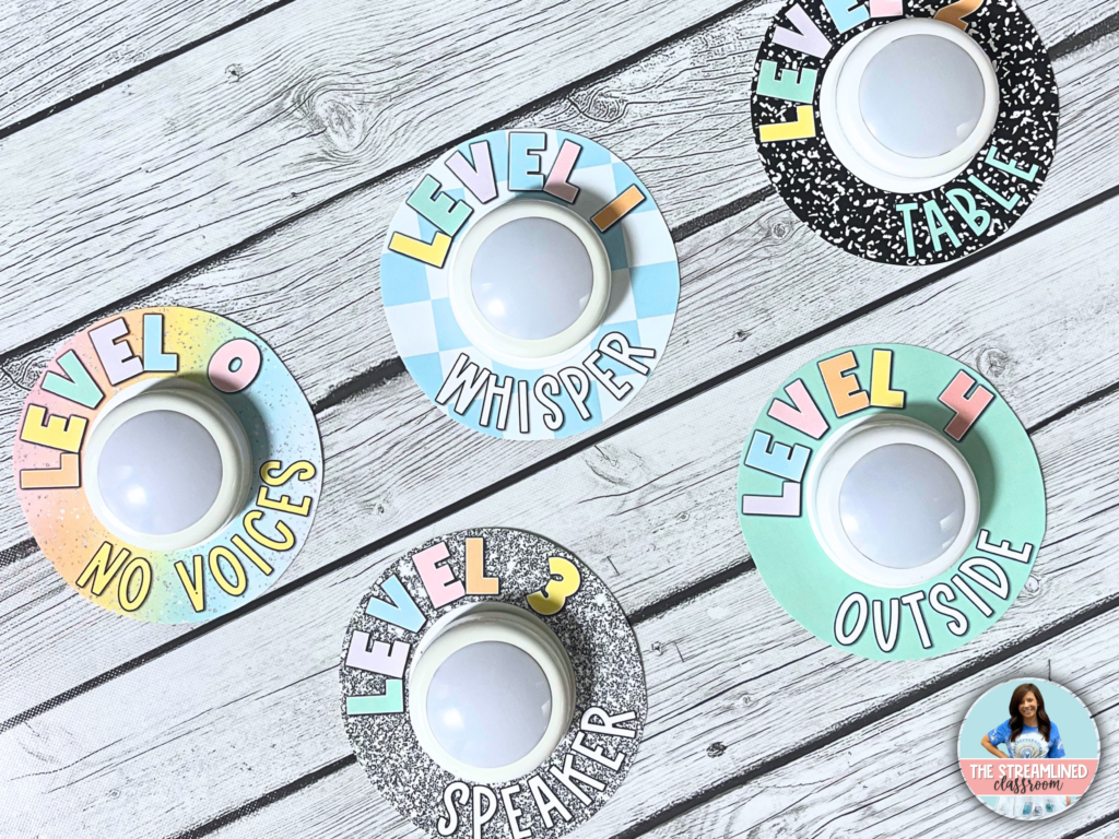 A set of colorful, composition notebook-themed push light voice level signs displayed on a wooden surface. Each sign features a different voice level, including "No Voices," "Whisper," "Speaker," "Table," and "Outside." The signs have a mix of pastel and black-and-white designs, making them a fun and functional classroom management tool.