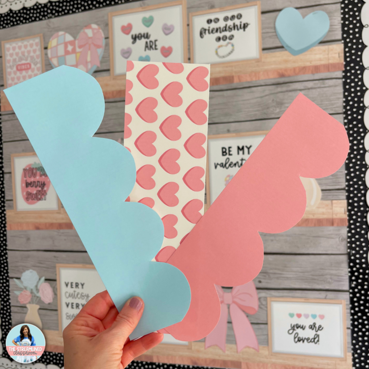 A teacher holding three different printable bulletin board border strips, featuring pastel scallops, pink candy hearts, and a soft blue design to complement the Valentine’s decor.
