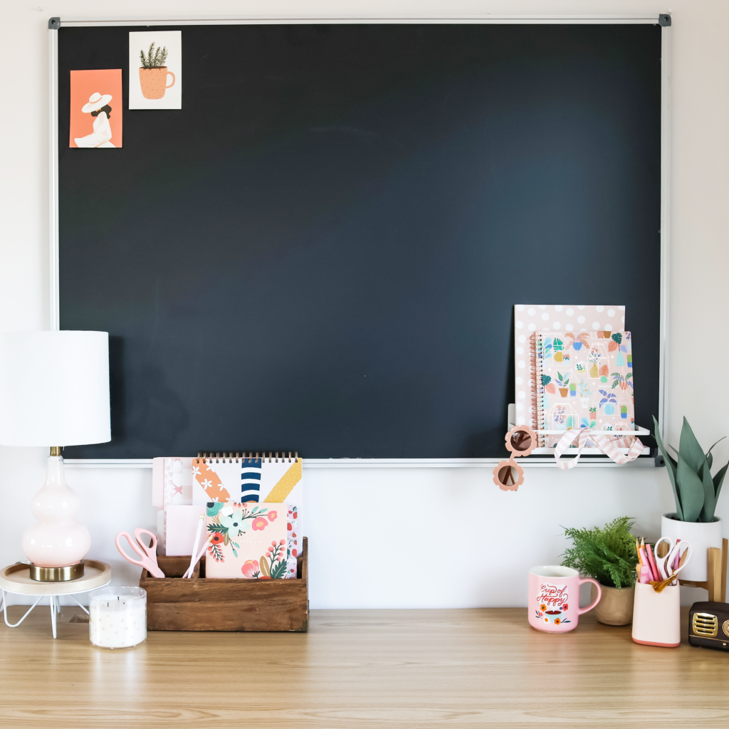 A neatly organized teacher's desk with a black bulletin board above it. The desk features a wooden organizer filled with colorful notebooks, a pastel pink lamp, a candle, and various office supplies in soft pink and neutral tones. On the board’s ledge, there are more notebooks, a pair of sunglasses, and decorative ribbon. Small plants and a motivational coffee mug with the words 'Simply Happy' add warmth to the setup, creating a cozy and stylish workspace.