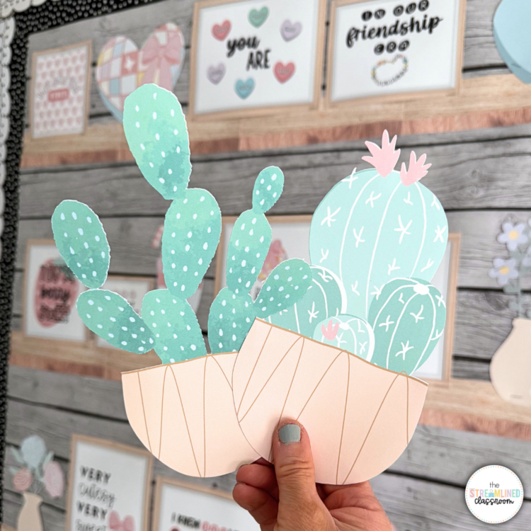 Close-up of a hand holding two cut-out cactus accents in pastel tones, with a boho classroom bulletin board in the background. Features soft neutral classroom decor elements and motivational quote posters.