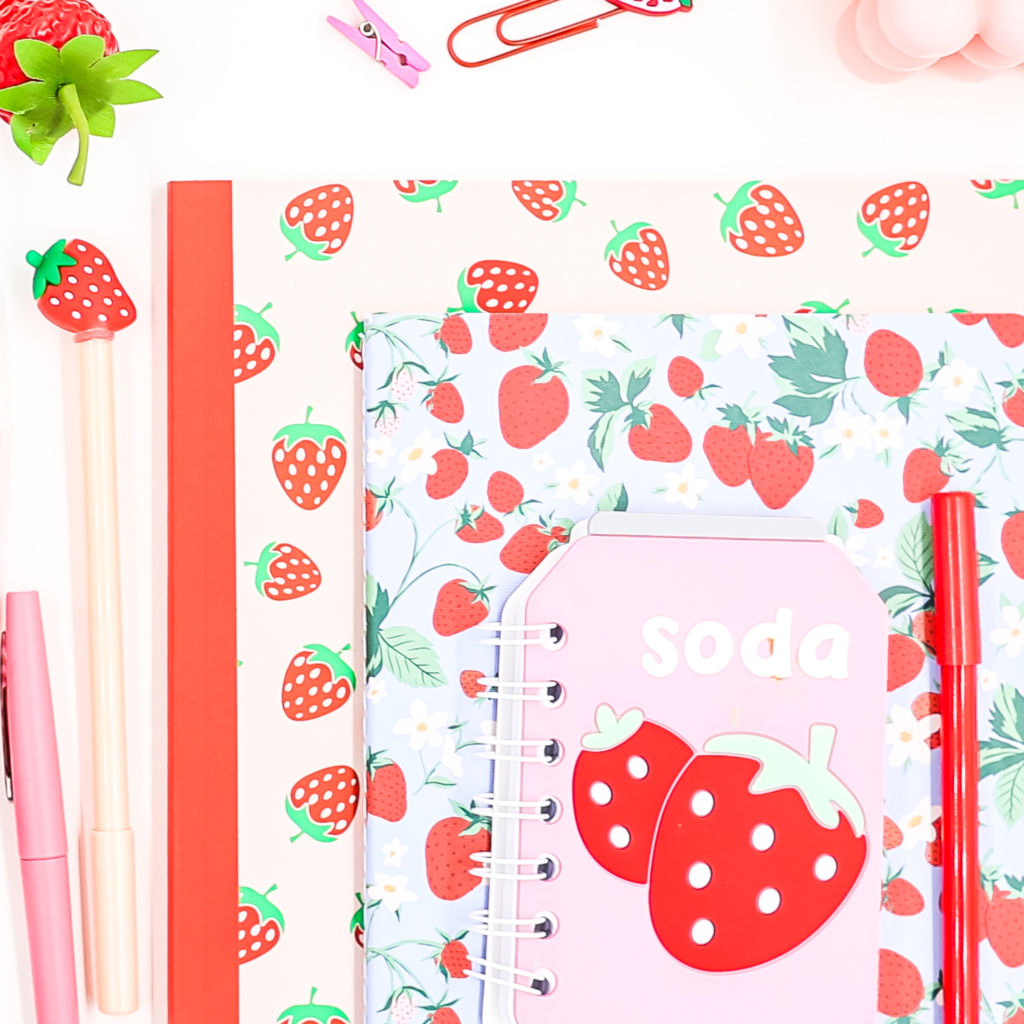 Flat lay of strawberry-themed stationery, including notebooks, pens, and accessories, arranged on a white background. Aesthetic and colorful teacher desk organization inspiration.
