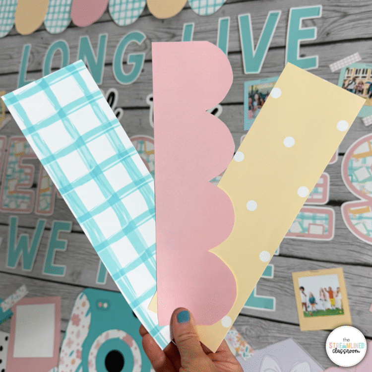 Three cut bulletin board border strips—blue plaid, pink scalloped, and yellow with white polka dots—held in hand in front of the decorated bulletin board.